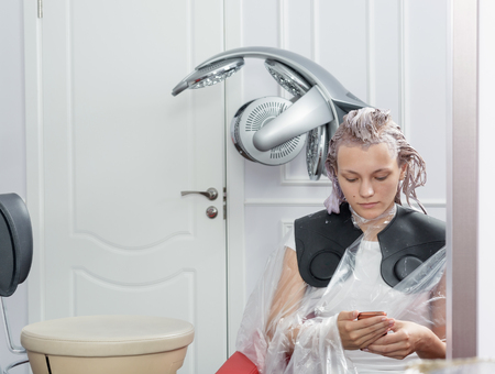 A blonde woman surfing the internet with her smartphone during hair colouringの写真素材