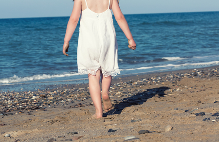 Little girl in white dress walking alone the sea, playing on the seashoreの写真素材