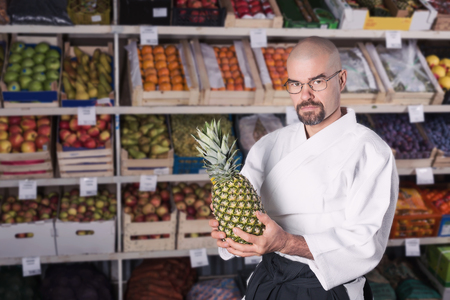 AVery hungry man against the background with racks with fruits and vegetables dressed in Japanese kimono and hakama holding a pineappleの写真素材
