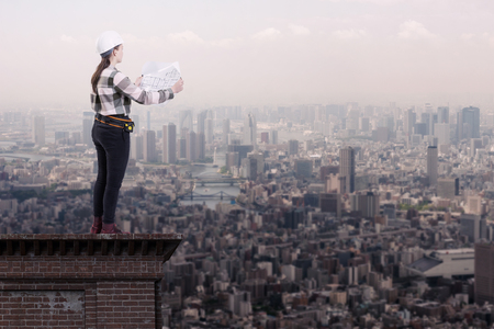 Female engineer holding construction plans standing on the roof top above a cityの写真素材