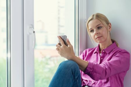 Pretty blonde girl sitting on a window and smartphone in handsの写真素材