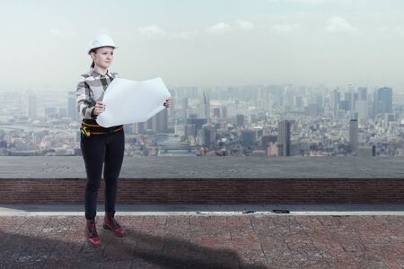 Female engineer holding construction plans standing on the roof top above a cityの写真素材