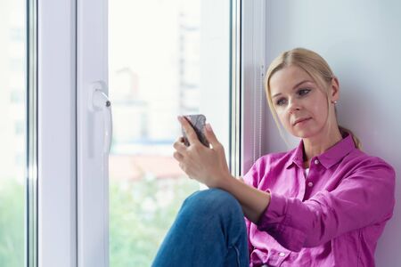 Pretty blonde girl sitting on a window with smartphone in handsの写真素材