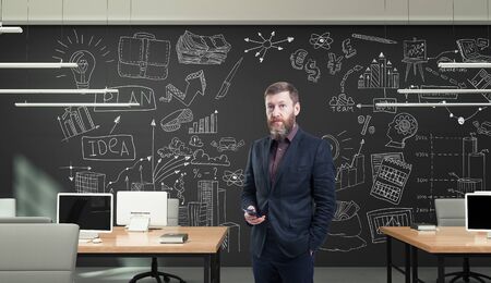 Bearded businessman in modern office holding smartphone in hand standing against a creative business strategy sketch drawn on a wallの写真素材