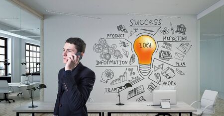 A young businessman talking on a phone standing in modern office with creative business strategy sketch drawn on white wallの写真素材