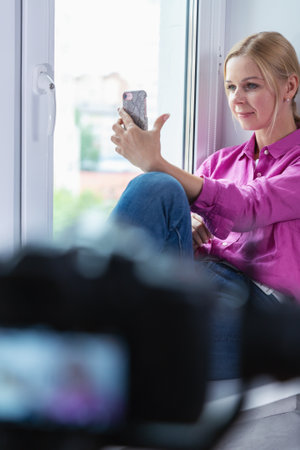 Pretty blonde girl sitting on a window with smartphone in handsの写真素材