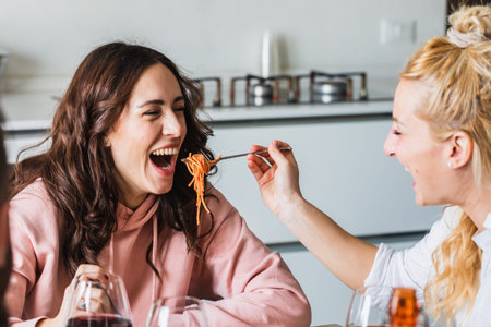 Couple of young girls friends laughing having fun joking with spaghetti. Blonde girl feeding her...