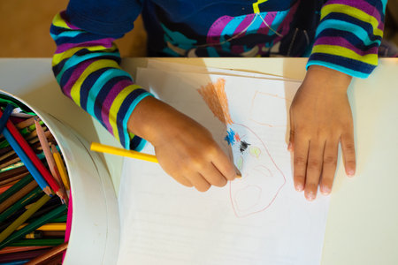 Closeup of child's hands while he is drawing, dressed in bright colors near a box of colorful crayonsの写真素材