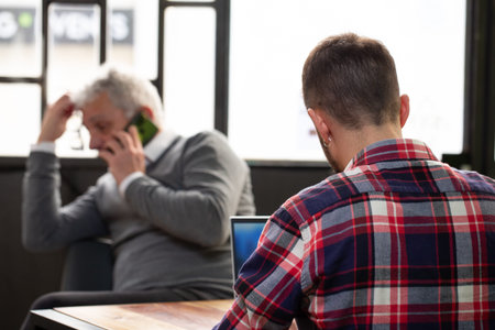 Anonymous situation with an unrecognizable young boy, with his back turned while using the laptop and an out of focus elderly gentleman phoning with his smartphone and glasses in his handの写真素材