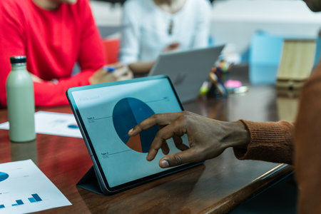 Hand of a dark-skinned man touching a digital tablet showing a pie chart on the screen. Office desk with several people working. Laptops, water bottle ad documents on the table. Co-working space.の写真素材
