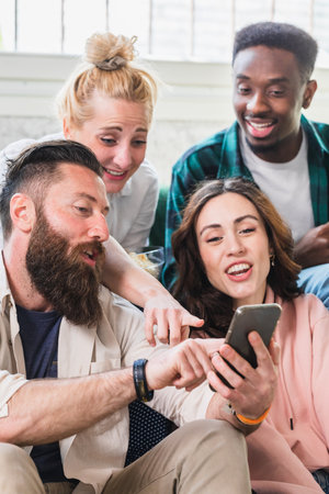Young friends of mixed nationalities laughing commenting on social media posts with smatphone. Jokes and laughter in a moment of leisure at homeの写真素材