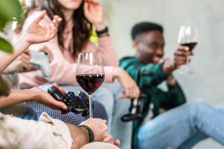 Selective focus on a boy's hand holding a goblet of red wine. Multiethnic party among friends with unrecognizable people. Console for gaming in one hand. Concept of having fun and getting high with alcoholの写真素材