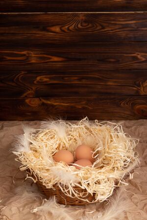 Chicken eggs lie on a wicked paper straw in a wicker basket on a dark wooden background in the morning sunの写真素材