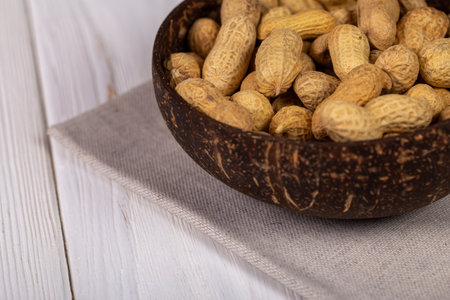 Close-up view of whole peanuts in their shells served in a natural, dark bowl, placed on a beige linen napkin on a white wooden tableの写真素材