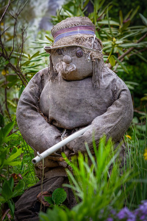 Front view of a homemade, rustic scarecrow made of burlap sack and straw, wearing a hat, holding a staff, and standing among lush green garden foliageの写真素材
