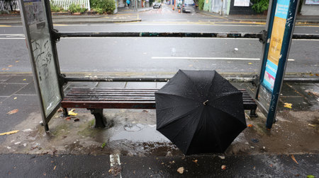 A lone umbrella left on a rainy bench at a bus stop, water pooling on the groundの素材