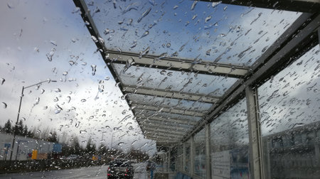 Gray skies above a bus stop while raindrops scatter across the shelter's plastic roofingの素材