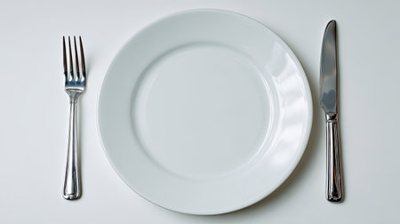 A minimalist composition featuring an empty white plate accompanied by a knife and fork, symbolizing readiness for a culinary experience. Ideal for food-related themes.の素材