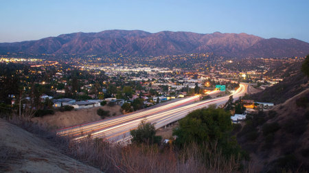 Captivating twilight view of a bustling city nestled among mountains, featuring illuminated traffic trails on the highway and serene natural surroundings.の素材