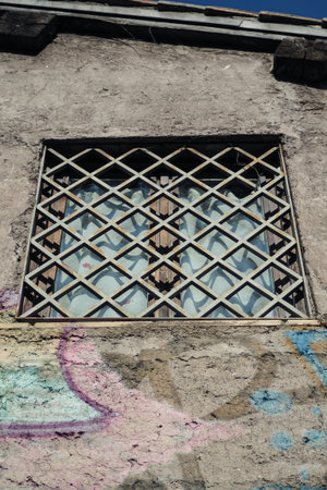Detail of window with criss cross shutter, in a shabby concrete wall with faded graffiti