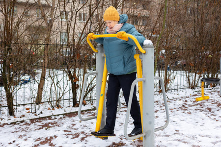 Activity. Mature woman in warm jacket and hat training on outdoor elliptical machine in snowy...