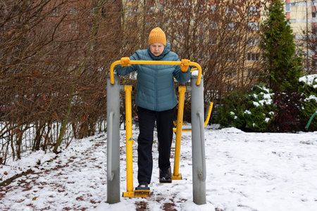 Activity. Mature woman in warm jacket and hat training on outdoor elliptical machine in snowy...