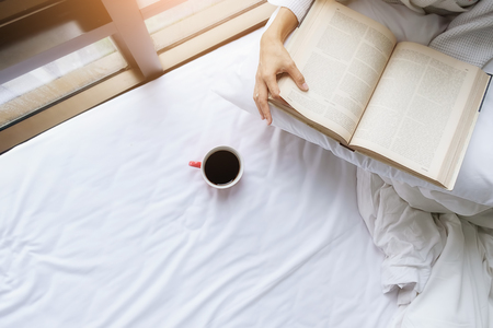 Reading a book. Beautiful female sitting on the bed near window and reading a book in her hands. Morning with a book in their hands. Relaxing concept. Vintage color,selective focus.の写真素材