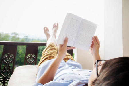 Rear view of asian man relaxing on a sofa and holding book on bed at home terrace with beautiful green background view. Relaxing concept.の写真素材