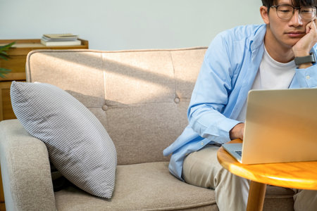 Young man sitting on sofa and using laptop in living room at homeの写真素材
