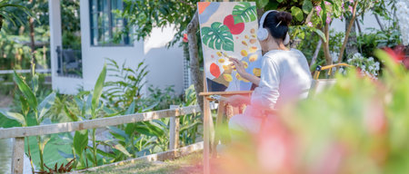 Asian woman painting on the wall in the garden. She is wearing white shirt and headphone.の写真素材