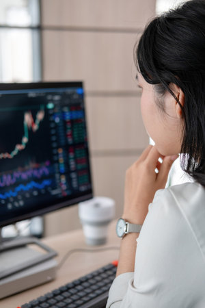 A young woman stand at her office desk, surrounded by a computer and tablet. She intently looks at...