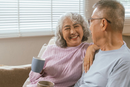 Happy couple spending quality time together in cozy modern home. They are having a conversation, drinking coffee while the wife is sitting on a sofa and husband on chair in the living room, displaying love, affection, togetherness.の写真素材