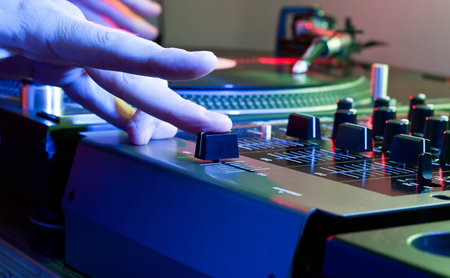 Hand of a DJ pushes the crossfader of a mixing desk in a colourful club environment