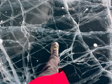 A foot of tourist standing on the cracks surface of frozen lake Baikal in the winter season of Siberia, Russia. Traveler man foot standing on cracks surface of the natural ice in frozen water. Top angle view of feet standing on broken iceの写真素材
