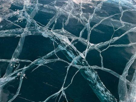 Abstract background of ice and cracks on the surface of frozen Lake Baikal. Spiderweb of cracks on slippery, smooth ice surface of a Baikal lake in Siberia (Russia). Cracks in the ice. Cracked ice on a frozen lake. The natural texture of winter ice with white bubbles and cracks on a frozen lake. Abstract bright background, flat layout, layout, top view. background. selective focus. Transparent dark blue ice of frozen Baikal lake with white cracks pattern. beautiful winter backgroundの写真素材
