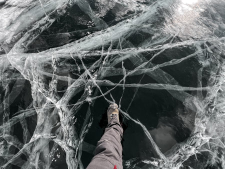 A foot of tourist standing on the cracks surface of frozen lake Baikal in the winter season of Siberia, Russia. Traveler man foot standing on cracks surface of the natural ice in frozen water. Top angle view of feet standing on broken iceの写真素材