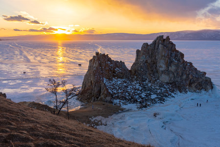 A group of people are walking on a frozen lake near a large rock. The sky is cloudy and the sun is setting, creating a moody atmosphere. View of Mount Shamanka, Lake Baikal in winterの写真素材