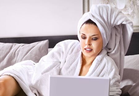 Rich woman in white bathrobe on a bed with laptopの写真素材