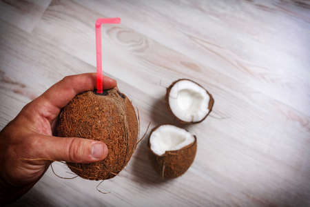 The adult man's hand holds a whole hairy coconut with an inserted straw over a light table close up. On the table are two cracked halves of coconut.の写真素材