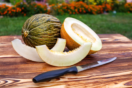 A whole and cut in half and sliced ripe melons lie on the wooden table, next to them a knife in...