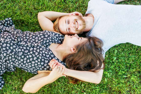 The beautiful couple white girl and her boyfriend lie on the green grass, hug and smile. The guy looks directly at the camera, the girl looks at him from the side. Bright sunny summer day, top view.の写真素材