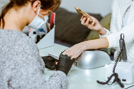 A young white manicurist girl makes a manicure to the client in the workplace. On her face a protective dressing, on her hands protective gloves. Close view.の写真素材