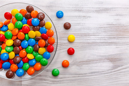 On a light surface of a wooden table there is a transparent round plate of mixed colored candy....