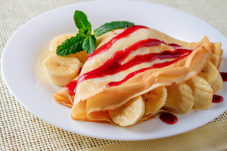 On the white plate with sliced ??bananas wrapped in pancakes. Pancakes are poured with jam and decorated with green mint leaves.の写真素材
