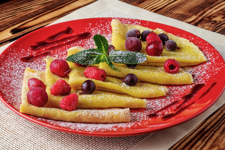 On the wooden table is a red plate. On it pancakes with berries and jam, sprinkled with powdered sugar and decorated with green mint leaves.の写真素材