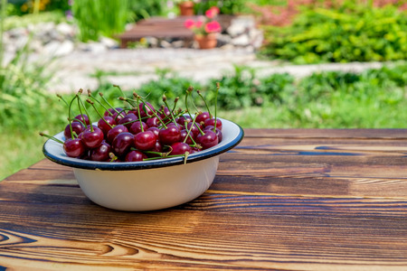 Many ripe red cherries in white metal bowl on the wooden table on open air. Close-up viewの写真素材