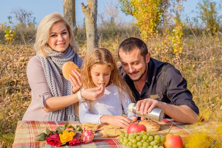 Mother and father feed daughter on picnic in autumn park. Sunny cloudless day. The concept of a happy family and carefree childhoodの写真素材