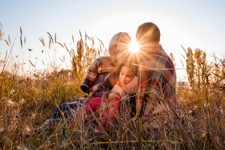 A little girl is sleeping in the arms of her parents who are sitting on the grass and covered with a plaid. Lens flare as element of composition. Concept of happy, reliability and carefree family lifeの写真素材