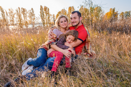 Bearded father hugs his wife and daughter. The young smiling family is sitting on the autumn grass and cover with a plaid. The little girl smiles with closed eyes. Happy family conceptの写真素材