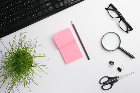 White table with keyboard, glasses, magnifying glass, pink stickers, clips, scissors, office plant and pencil. Minimalist workplace. Flat lay, copy spaceの写真素材
